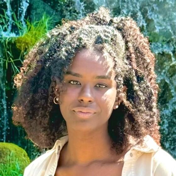 A headshot of a young woman with voluminous, dark curly hair, some of which is pulled back at the crown. She is looking at the camera with a soft, neutral expression and is wearing a light-colored, button-down shirt and small hoop earrings. The background is a bright, lush outdoor setting.