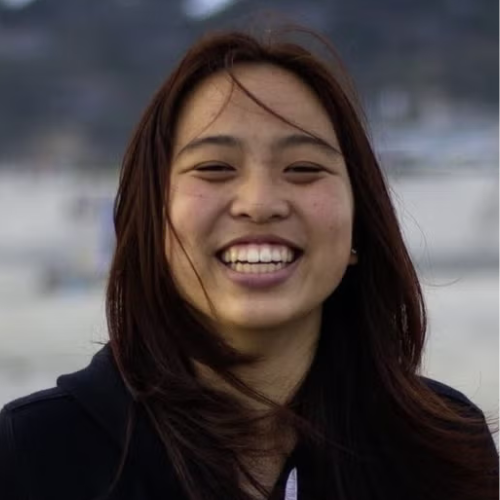 A close-up headshot of a young woman with long, dark hair blowing slightly in the wind. She is laughing joyfully at the camera, showing a wide, bright smile. She is wearing a dark-colored top. The background is a soft-focus, outdoor setting that appears to be a beach or a coastal area under overcast light.