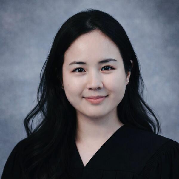 A headshot of a young woman with long, dark, wavy hair. She is wearing a black v-neck garment