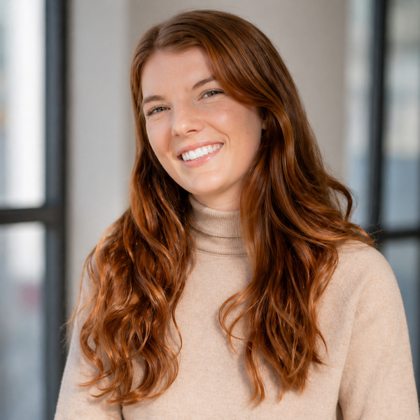 A professional headshot of a smiling woman with long, wavy auburn hair. She is wearing a light beige turtleneck sweater and is angled slightly toward the camera against a softly blurred indoor background with large windows.