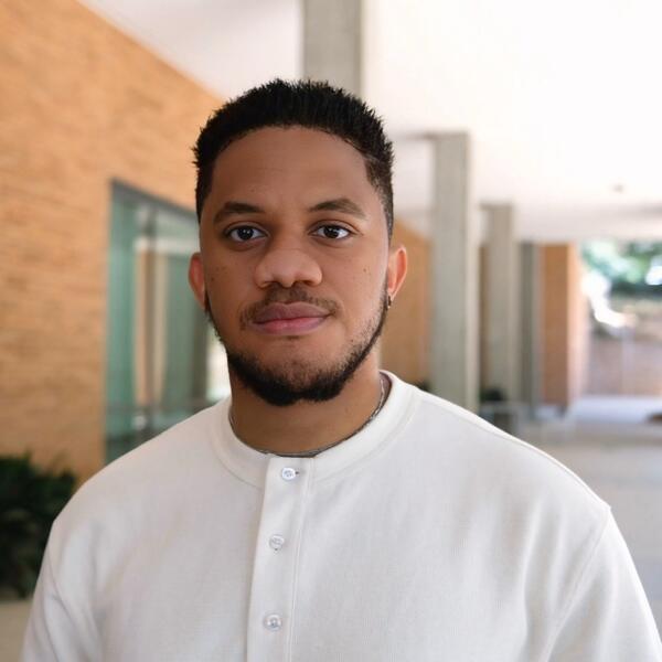 A headshot of a young man with short, dark hair. He is wearing a white, button-up shirt. He has a neutral expression and is looking directly at the camera. The background is a slightly blurred outdoor or campus setting with light brown brick walls and concrete pillars under bright, natural light.