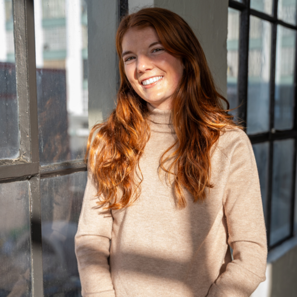 A medium shot of a young woman with long, wavy red hair smiling brightly. She is wearing a tan or beige turtleneck sweater. She is standing next to a large, industrial-style window with dark frames, through which soft sunlight is streaming and creating shadows across her sweater. The background shows a hint of an urban setting through the glass.