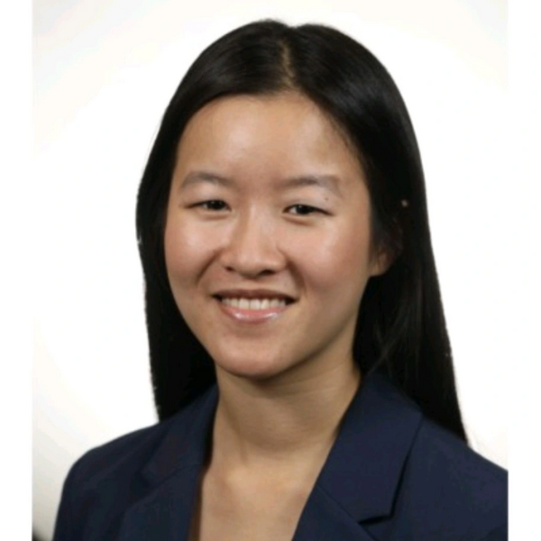  A headshot of a young woman with long, straight dark hair parted in the middle. She is smiling at the camera and wearing a professional navy blue blazer. The photo is set against a plain, bright white background with clean, studio-style lighting.
