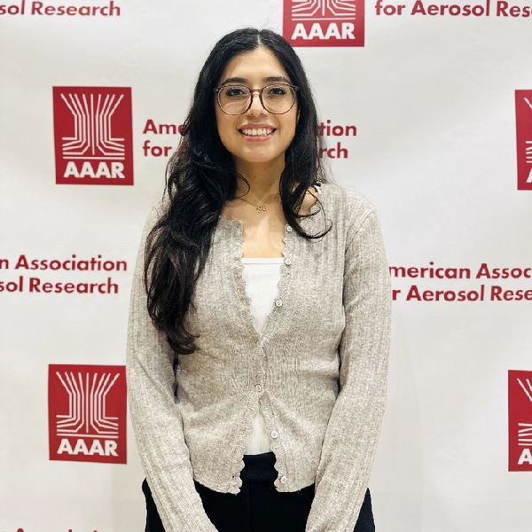 A medium shot of a young woman with long, wavy dark hair and glasses, smiling at the camera. She is wearing a light-colored, button-up cardigan over a white top and dark trousers. She stands in front of a white backdrop featuring the repeating red logo and text for the American Association for Aerosol Research (AAAR).